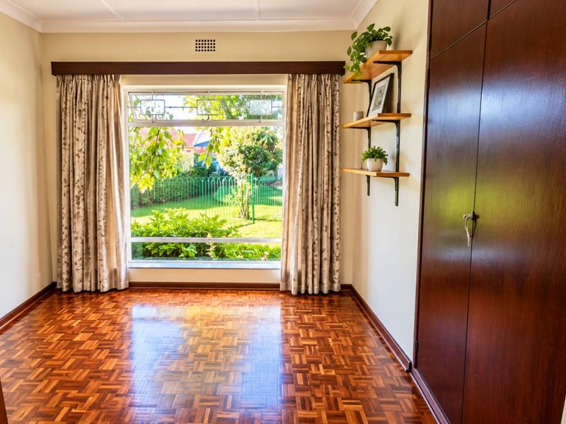 A bright, empty bedroom featuring polished parquet flooring, built-in wooden cupboards, and a large window overlooking the garden.