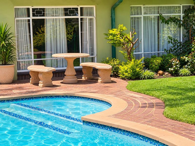 A well-maintained outdoor pool area featuring brick paving, a clear swimming pool, and direct access to the home through large windows with security bars.
