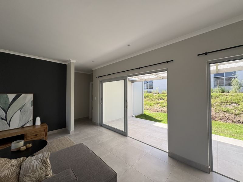 Modern living room featuring large sliding glass doors that open onto a covered patio, with neutral tiled flooring and a contemporary dark accent wall.