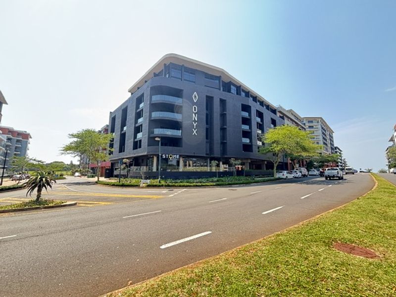 A modern multi-story apartment building featuring a dark facade, glass balconies, and prominent signage, situated on a corner lot with surrounding landscaped areas.