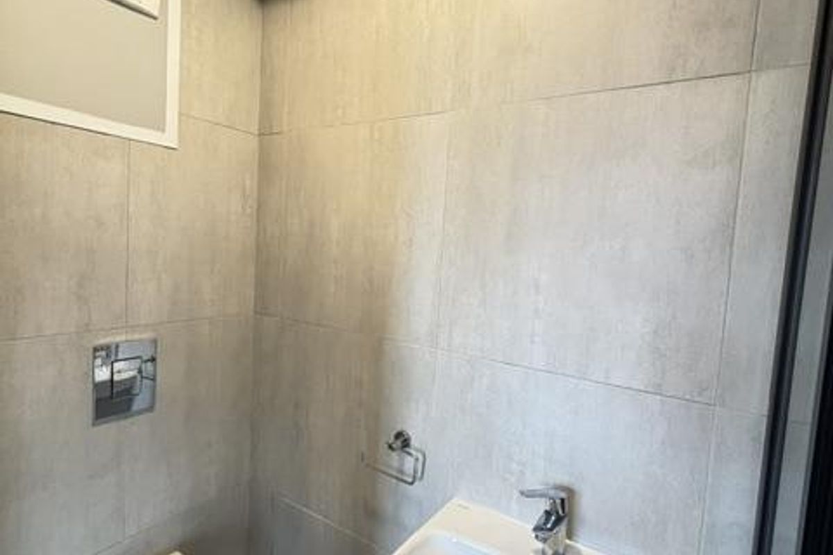 A modern, compact bathroom featuring a wall-mounted toilet, pedestal sink with chrome fixtures, and neutral floor-to-ceiling tiling.