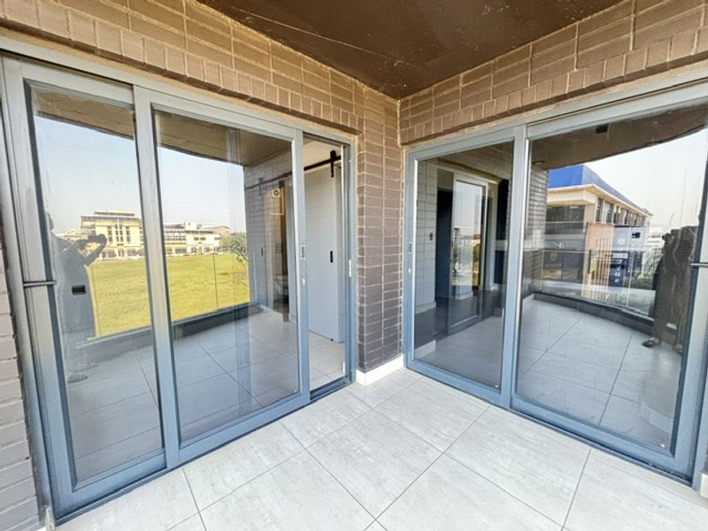A modern balcony featuring large sliding glass doors, tiled flooring, and a glass balustrade with views of the surrounding area.
