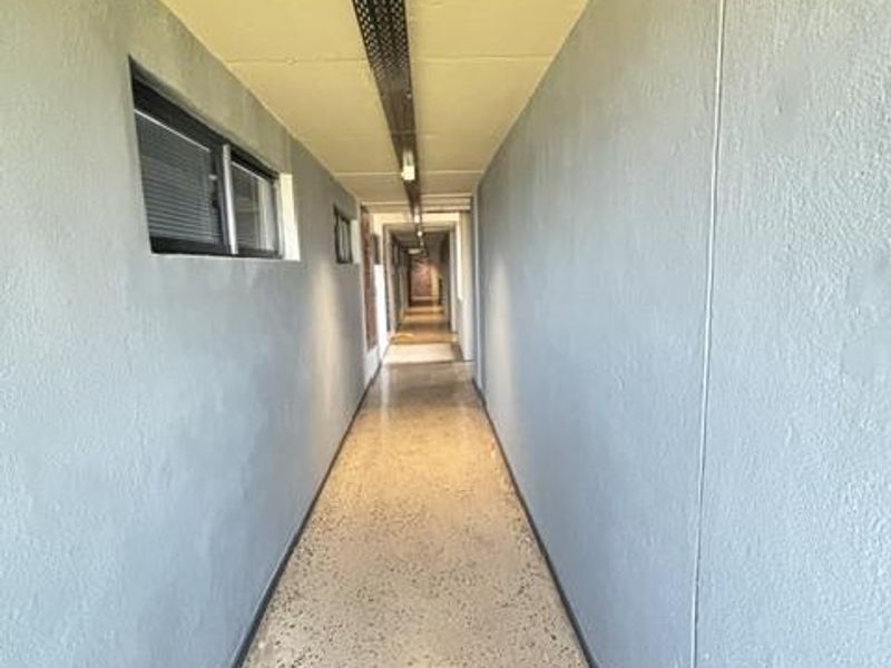 A long, utilitarian apartment hallway featuring terrazzo flooring, exposed cable management systems, and side-facing casement windows.