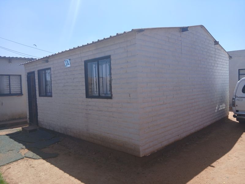 A modest single-story residential structure featuring painted brick walls, metal window frames with security bars, and a corrugated metal roof.