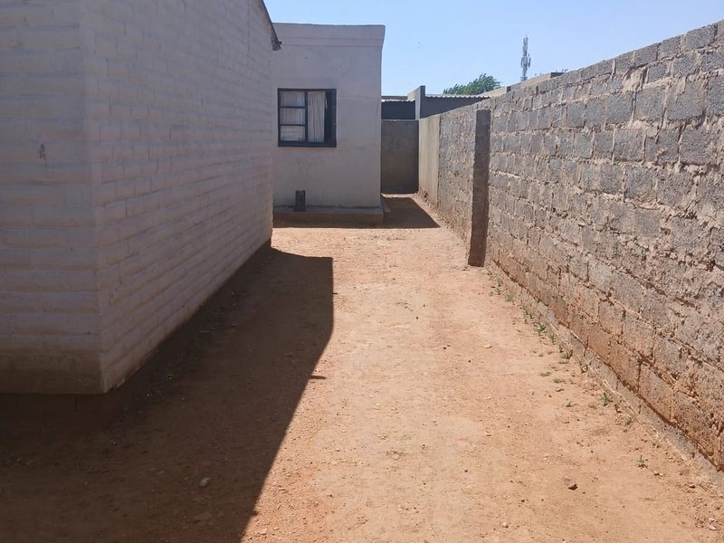 A narrow side passage between a painted brick building and a high masonry boundary wall with an unpaved dirt ground surface.