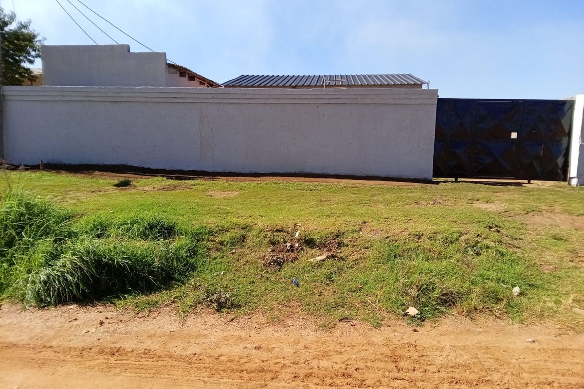 Front view of the property featuring a perimeter wall, metal gate, and corrugated roof structure with an unpaved verge area.