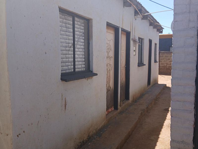 Exterior view of the rear building facade featuring a narrow concrete walkway, multiple access doors, and metal-framed windows with visible signs of weathering.