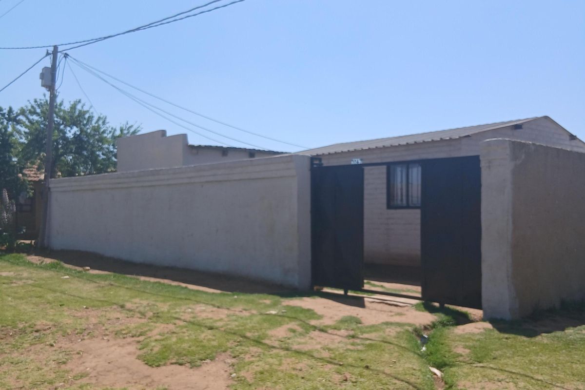 Front view of a property featuring a perimeter wall, metal gate entrance, and corrugated roof structure set on an unpaved plot.