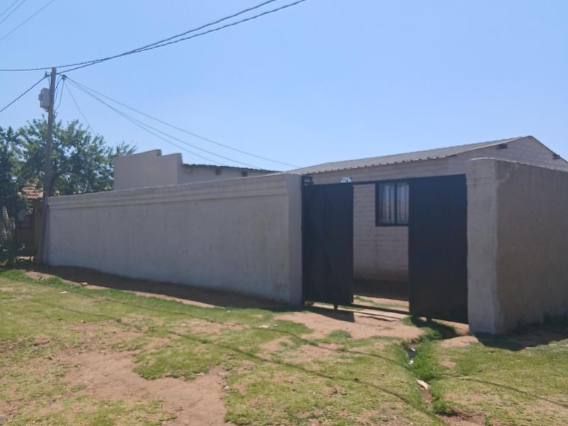 Front view of a property featuring a perimeter wall, metal gate entrance, and corrugated roof structure set on an unpaved plot.