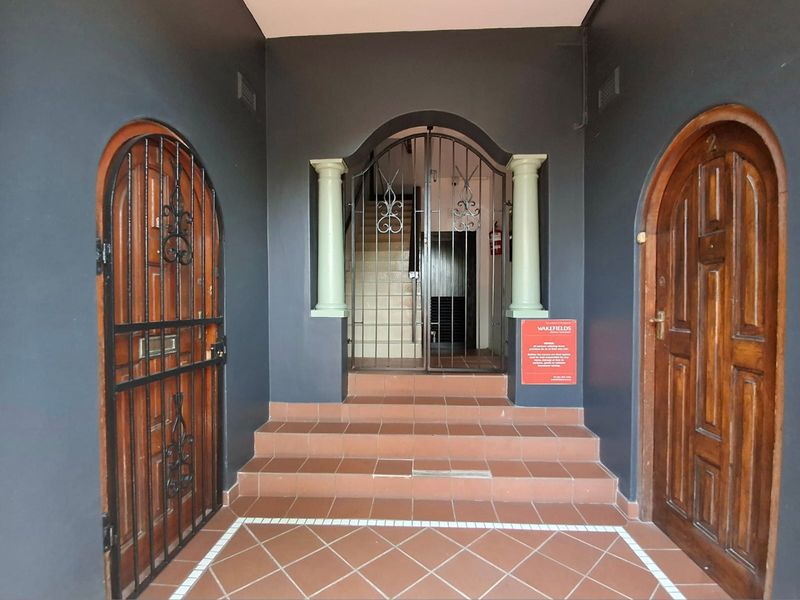 A secure apartment building entrance featuring arched wooden doors, decorative columns, and a tiled staircase leading to the upper levels.