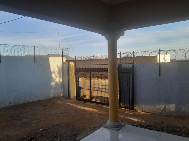 A covered patio area featuring a concrete column and tiled flooring, overlooking a secured perimeter wall with razor wire and a sliding gate.