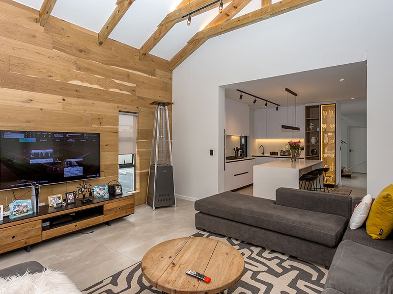 Modern open-plan living area featuring high vaulted ceilings with exposed timber trusses, wood-paneled feature wall, and a sleek, contemporary kitchen with an island.