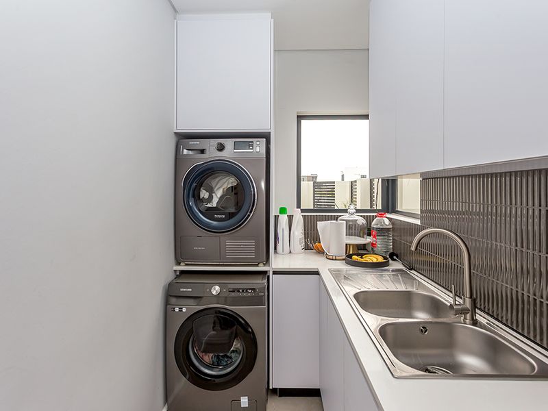 Modern laundry room featuring sleek white cabinetry, a stainless steel double sink with a gooseneck mixer, and a stacked washer and dryer setup.
