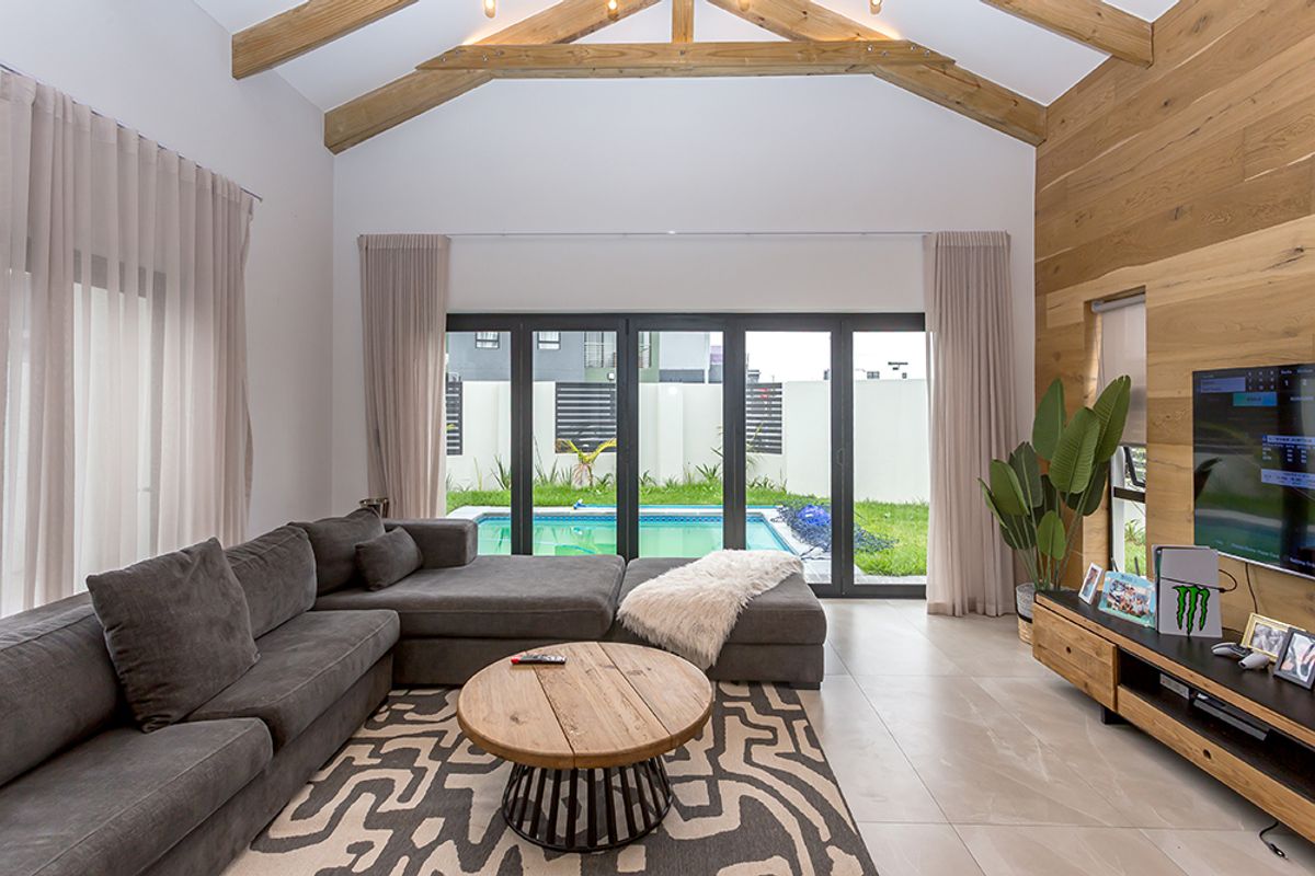 Spacious living room featuring a vaulted ceiling with exposed timber trusses, timber feature wall, tiled floors, and large sliding doors opening to a pool area.