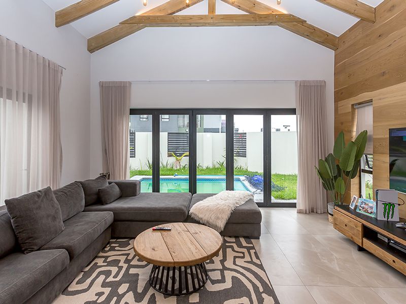 Spacious living room featuring a vaulted ceiling with exposed timber trusses, timber feature wall, tiled floors, and large sliding doors opening to a pool area.