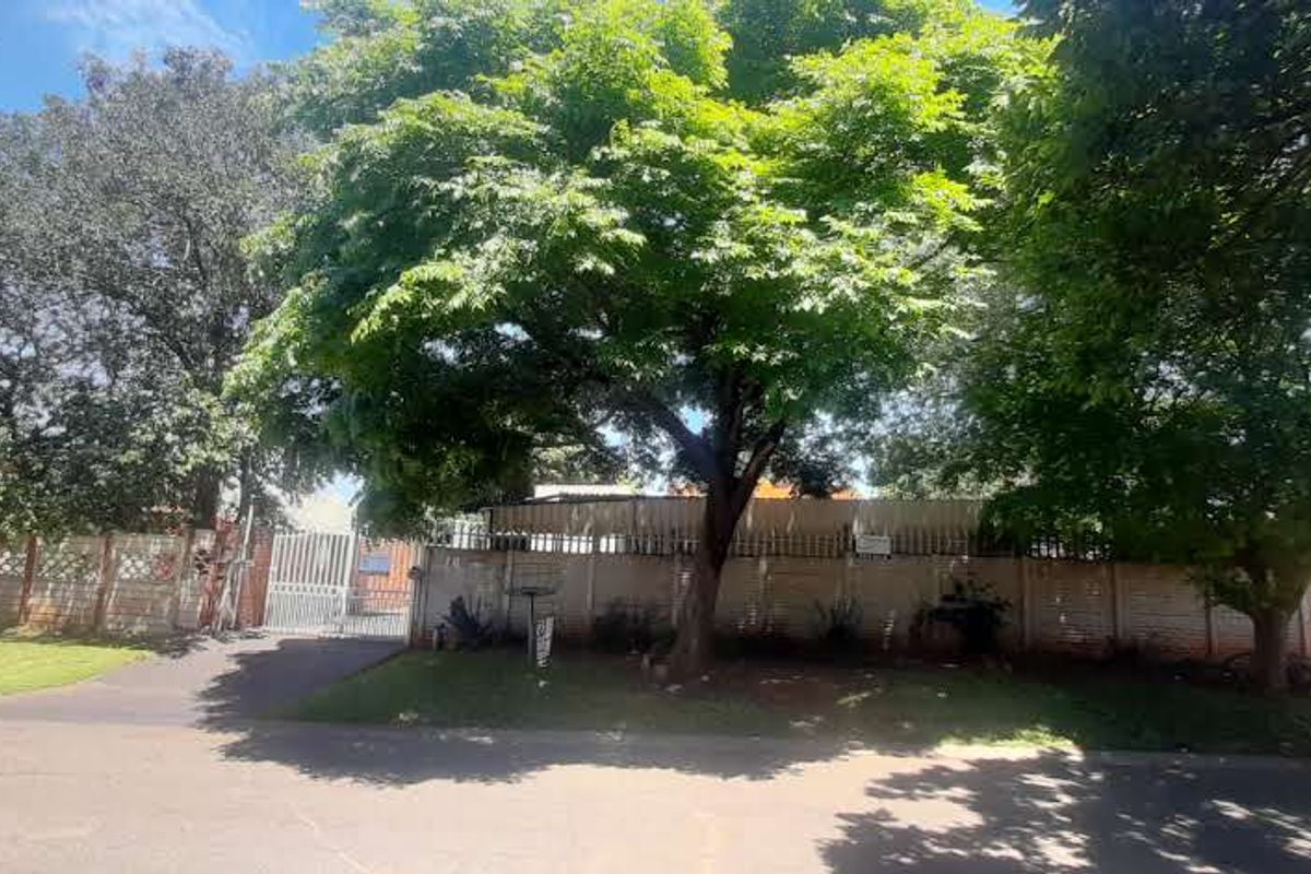Front view of the property featuring a secure metal gate, paved driveway, and a perimeter precast concrete boundary wall surrounded by mature trees.