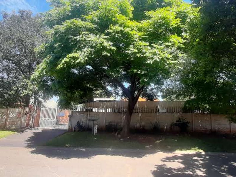 Front view of the property featuring a secure metal gate, paved driveway, and a perimeter precast concrete boundary wall surrounded by mature trees.
