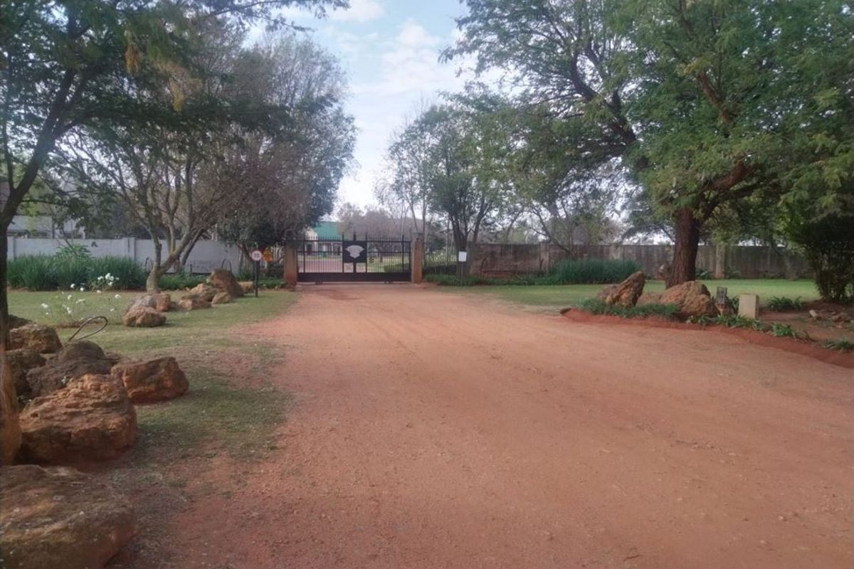 A view of the property entrance featuring a metal security gate, perimeter walling, and a landscaped garden area along an unpaved driveway.