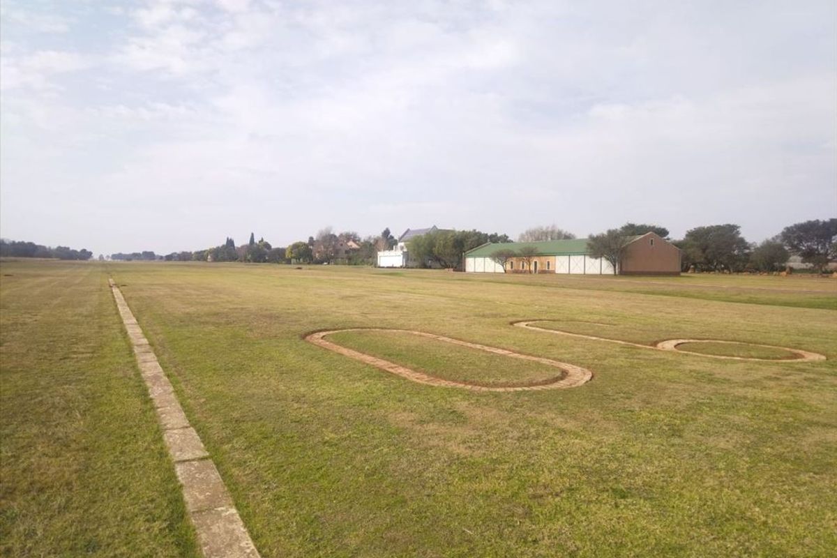 A wide, open residential land plot featuring a large grass lawn with decorative brick-edged circular features and distant outbuildings.