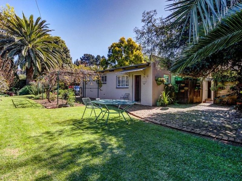 A rear exterior view of the property featuring a well-maintained lawn, a cobblestone patio area, and established garden landscaping with mature trees.