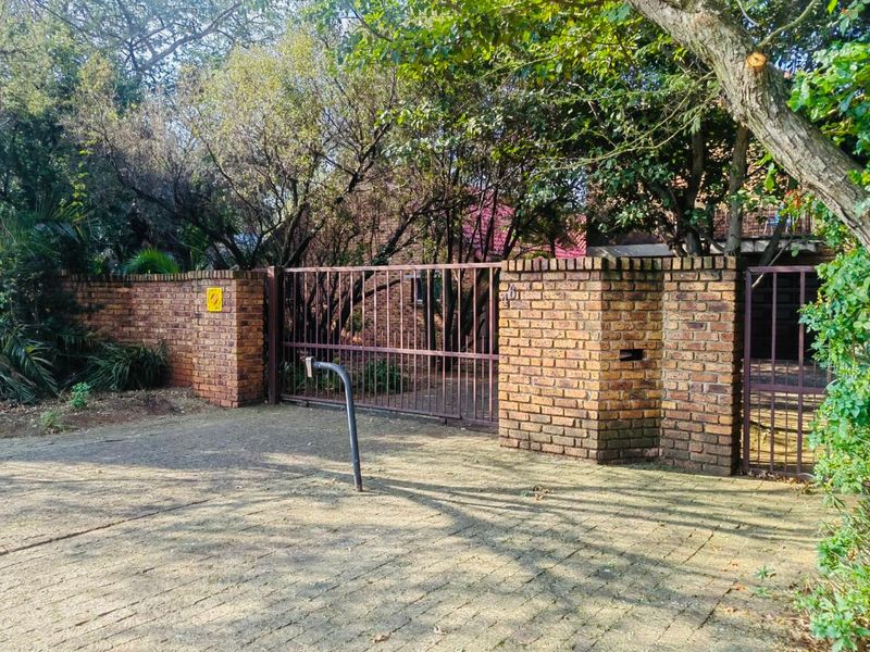 Front entrance featuring a paved driveway, brick boundary wall, and metal security gates, surrounded by mature trees and established garden greenery.