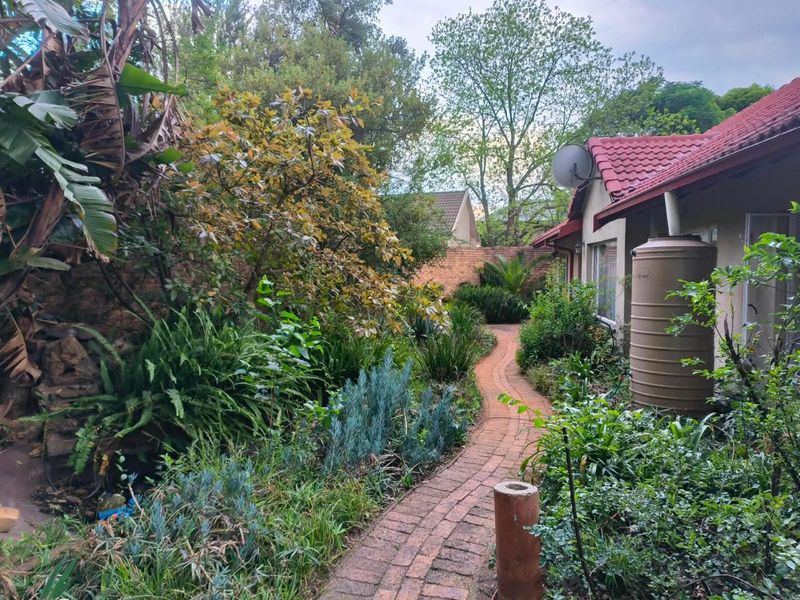 A lush, established garden featuring a winding brick pathway, mature greenery, and a functional water storage tank alongside the house exterior.