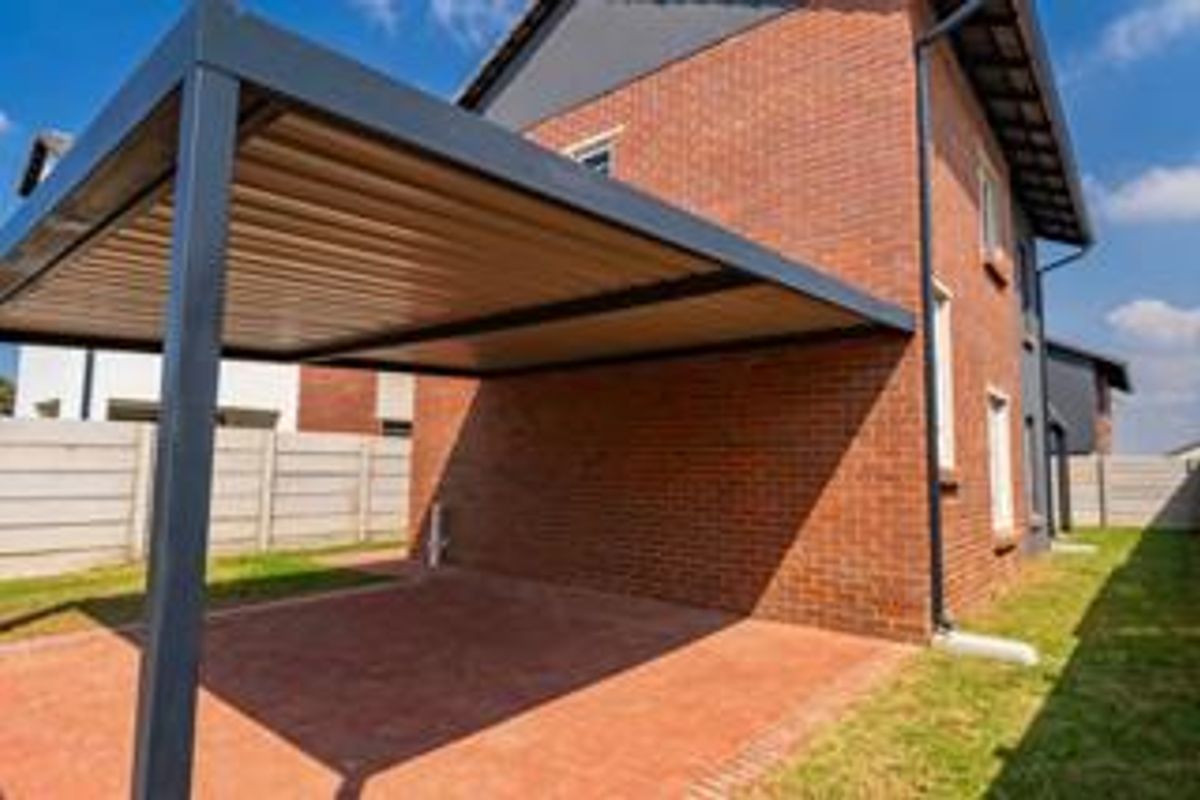 A well-maintained rear exterior view featuring a modern metal carport, red brick facade, and paved driveway area enclosed by a precast boundary wall.