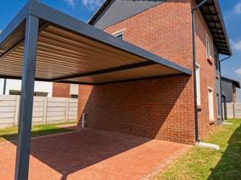 A well-maintained rear exterior view featuring a modern metal carport, red brick facade, and paved driveway area enclosed by a precast boundary wall.