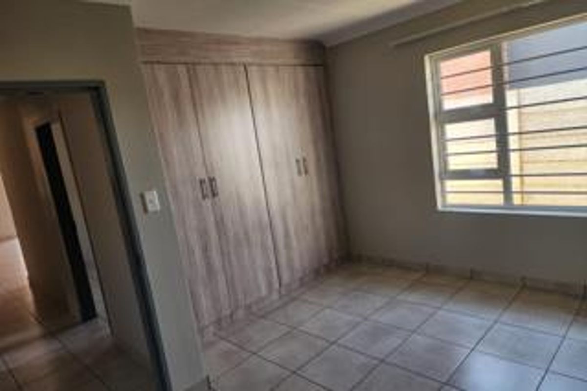 A clean, empty bedroom featuring neutral tiled flooring, built-in wooden cupboards, and a window fitted with security bars.