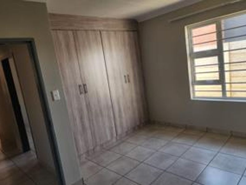 A clean, empty bedroom featuring neutral tiled flooring, built-in wooden cupboards, and a window fitted with security bars.