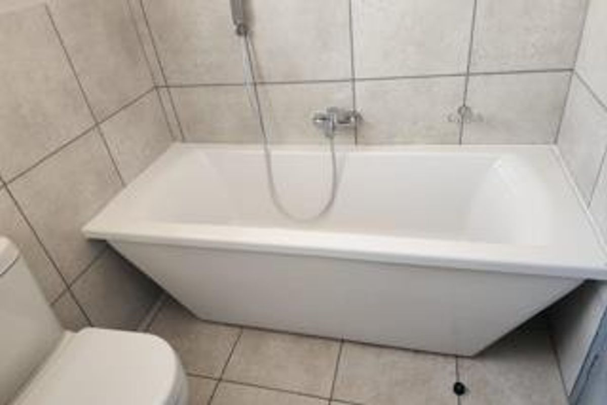 A clean, modern bathroom featuring a white bathtub with a wall-mounted shower mixer and neutral ceramic tiling throughout.