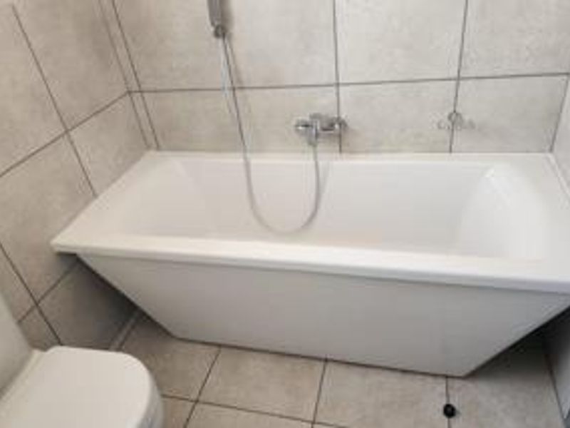 A clean, modern bathroom featuring a white bathtub with a wall-mounted shower mixer and neutral ceramic tiling throughout.