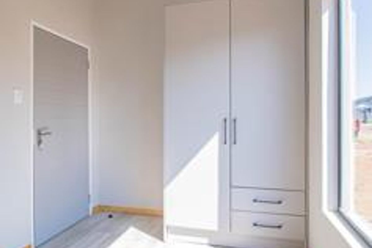 A bright, modern bedroom featuring light-toned laminate flooring, a built-in wardrobe with drawers, and a large window providing natural light.