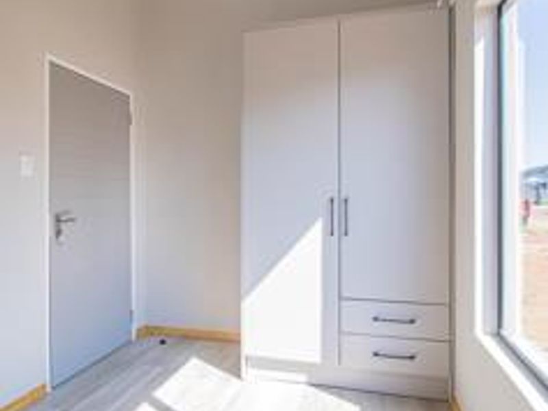 A bright, modern bedroom featuring light-toned laminate flooring, a built-in wardrobe with drawers, and a large window providing natural light.
