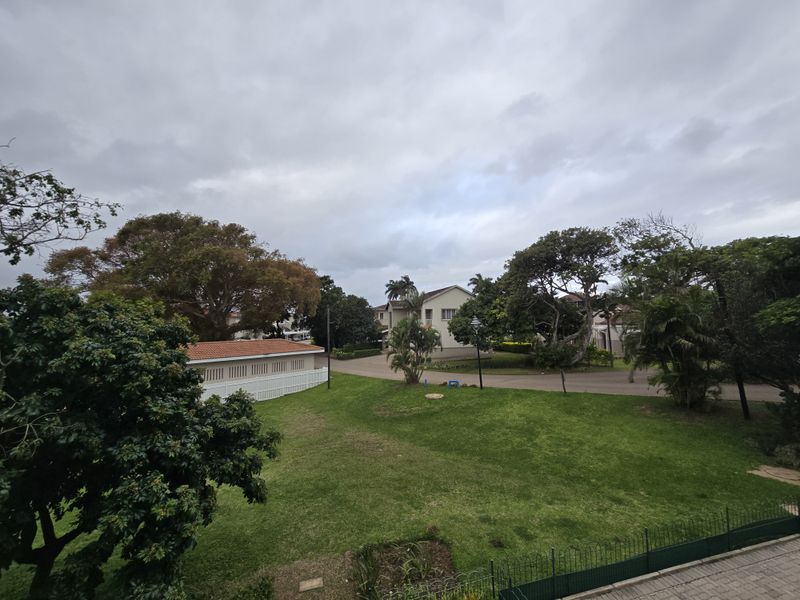 An elevated view overlooking a well-maintained communal garden area with mature trees, paved pathways, and surrounding residential buildings.
