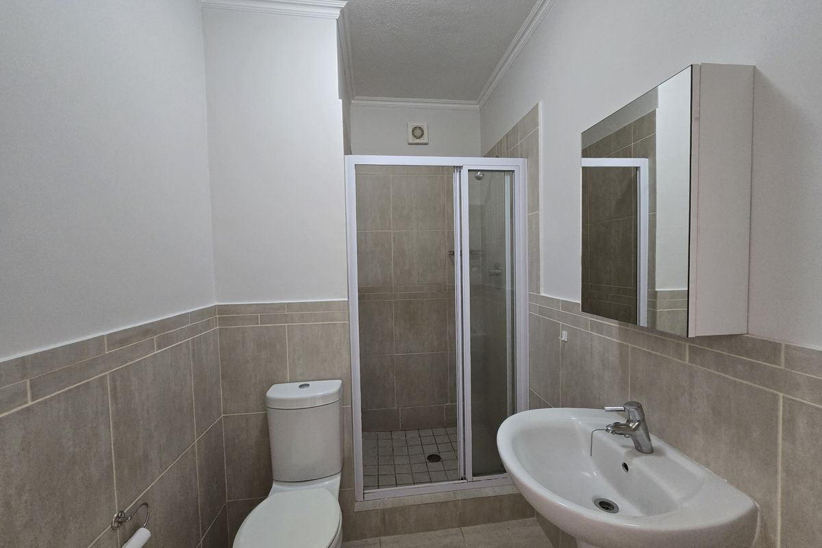 A clean and functional bathroom featuring a shower enclosure, pedestal basin, and wall-mounted medicine cabinet with neutral tiled finishes.