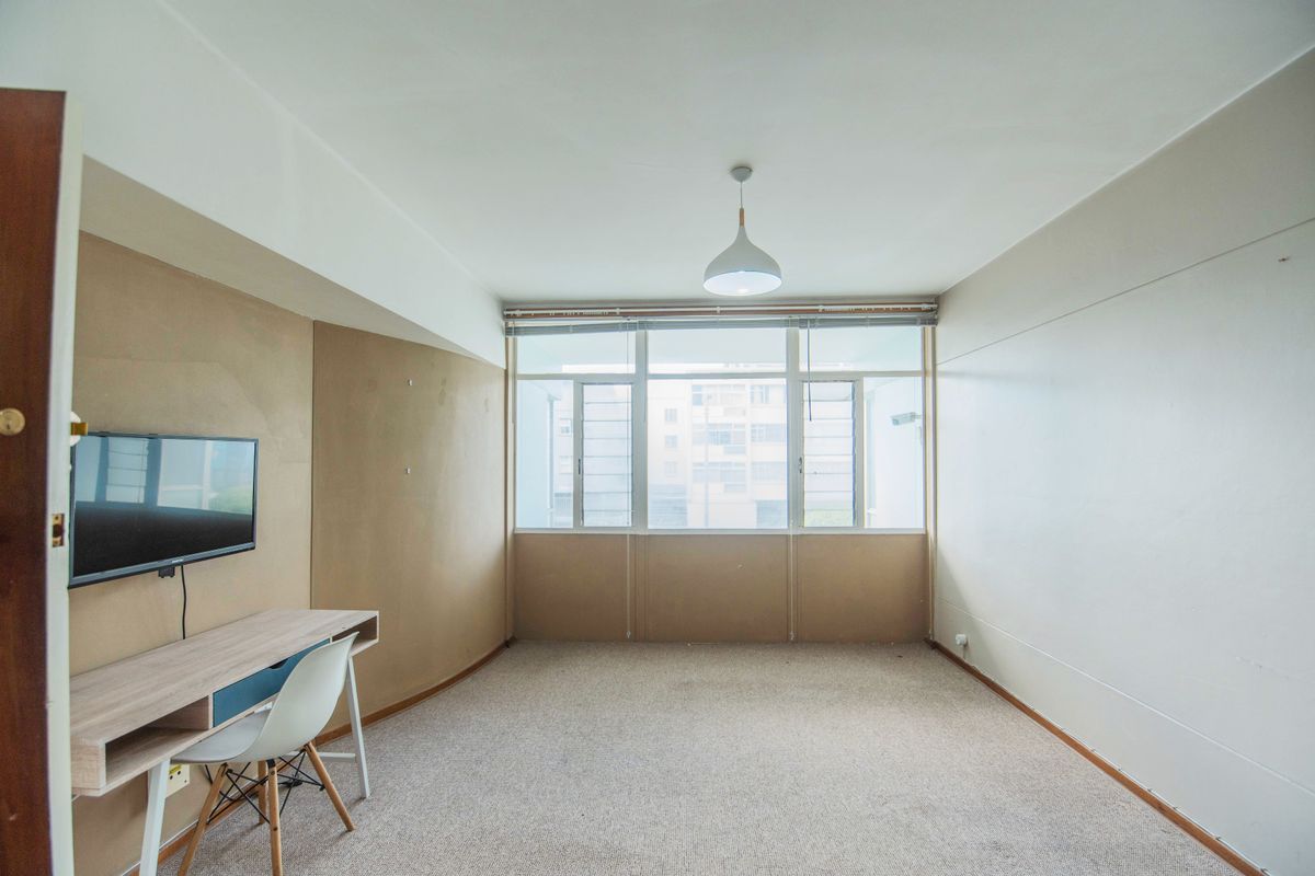 A spacious, carpeted bedroom featuring a large window that provides natural light and a central pendant light fixture.