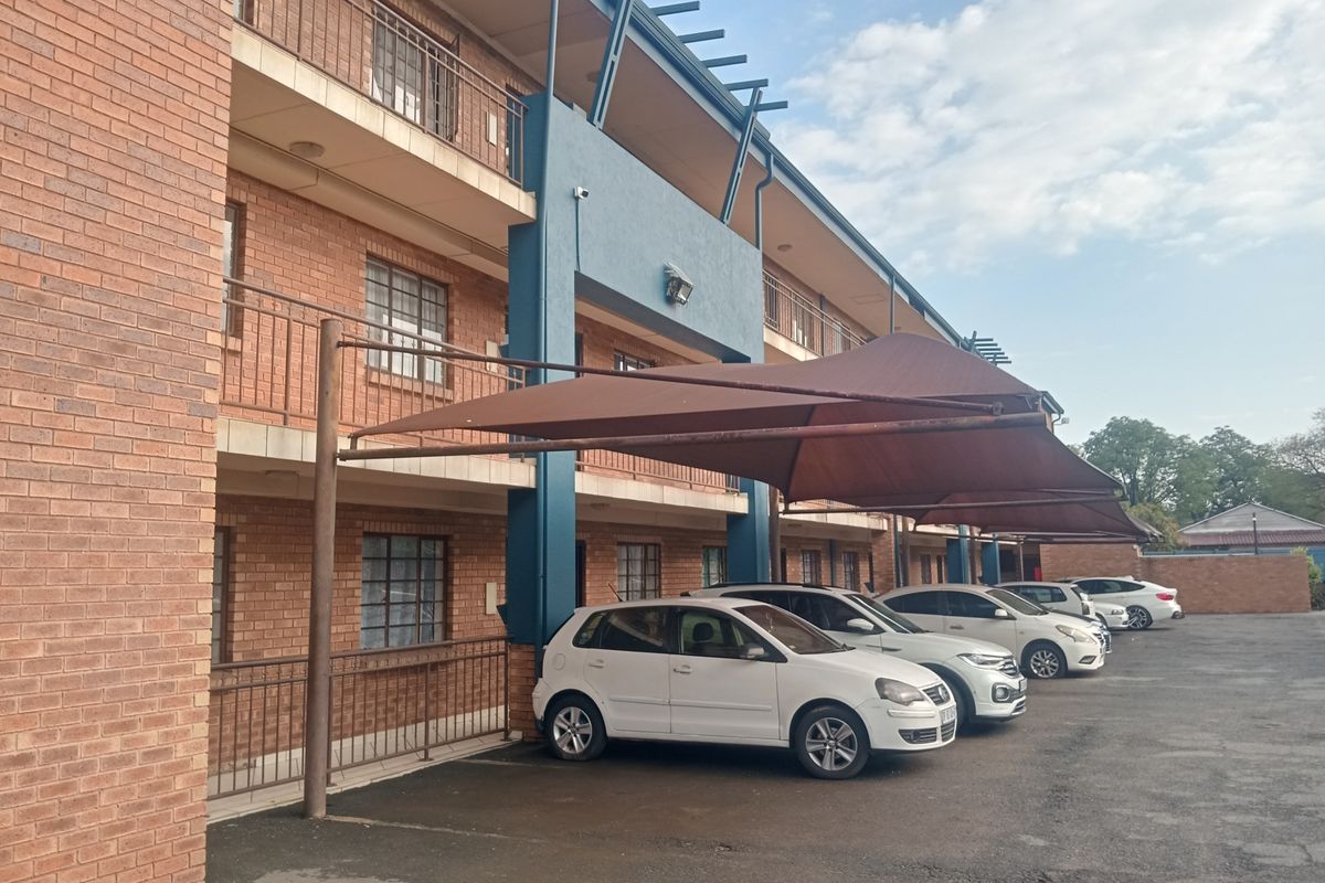 Exterior view of a multi-story apartment building featuring face brick facade, private balconies, and covered parking bays with shade cloth structures.