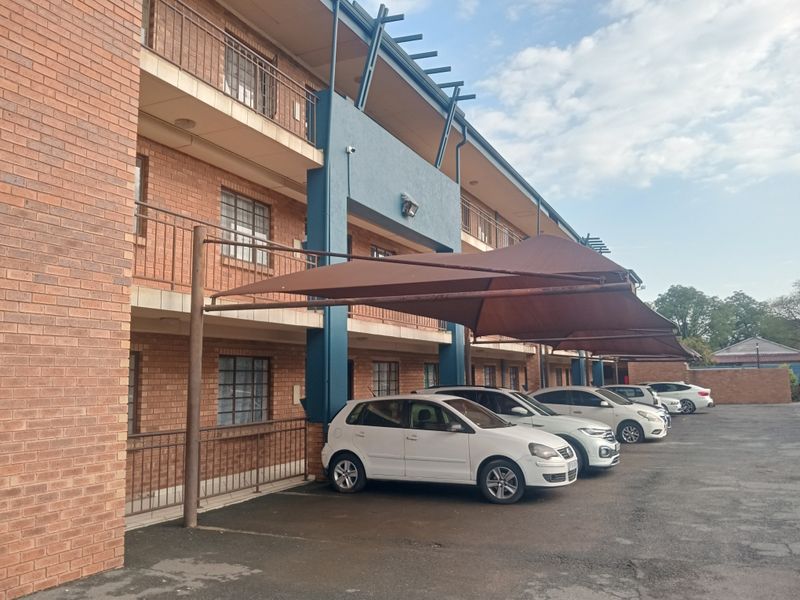 Exterior view of a multi-story apartment building featuring face brick facade, private balconies, and covered parking bays with shade cloth structures.