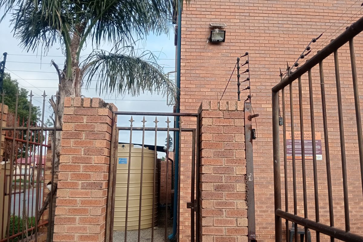 Rear exterior area featuring a brick perimeter wall, metal security gate with electric fencing, and a water storage tank.