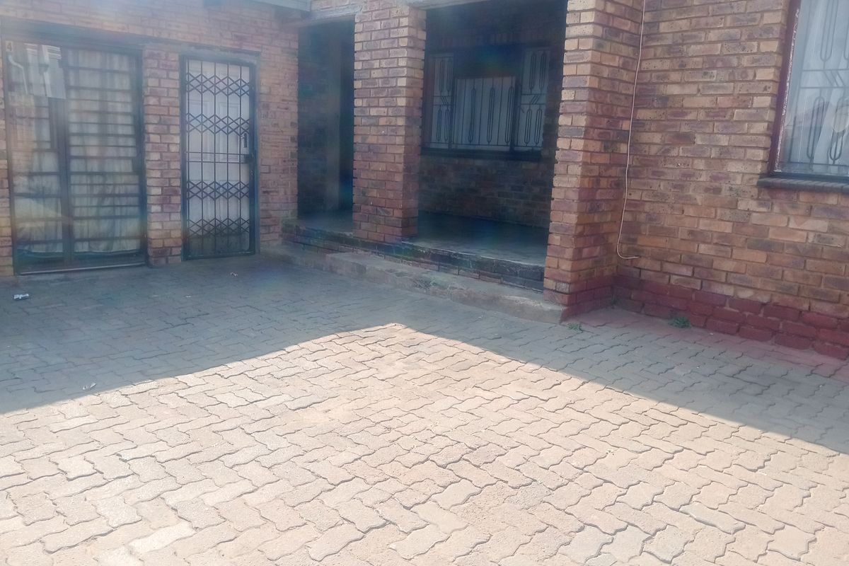 A rear exterior view of a face brick house featuring a paved courtyard area and a covered patio entrance with security gates.