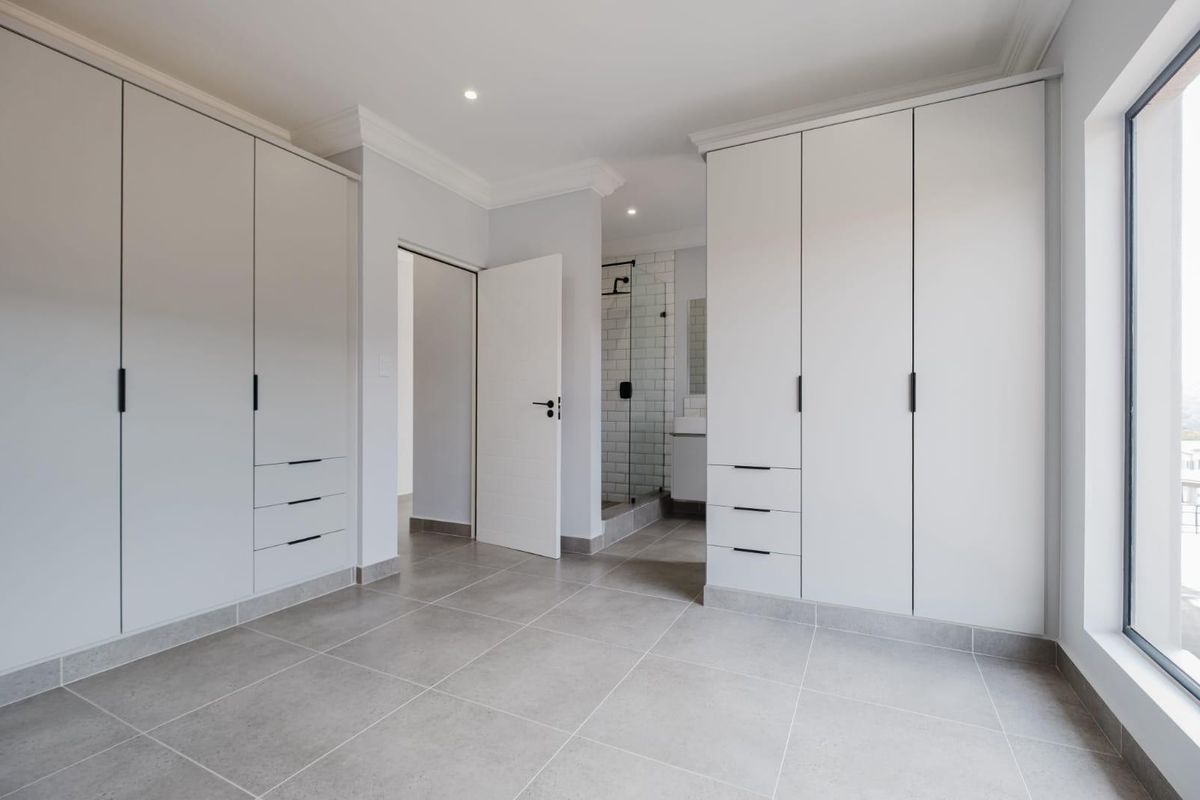 A bright, modern bedroom featuring extensive built-in cabinetry, neutral tiled flooring, and direct access to an en-suite bathroom.