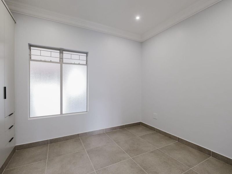 A clean, empty bedroom featuring neutral tiled flooring, a built-in cupboard, and a frosted glass window with modern finishes.
