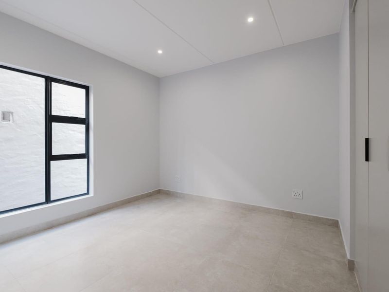 A clean, modern, and empty bedroom featuring neutral tiled flooring, built-in cupboards, and a large black-framed window providing natural light.