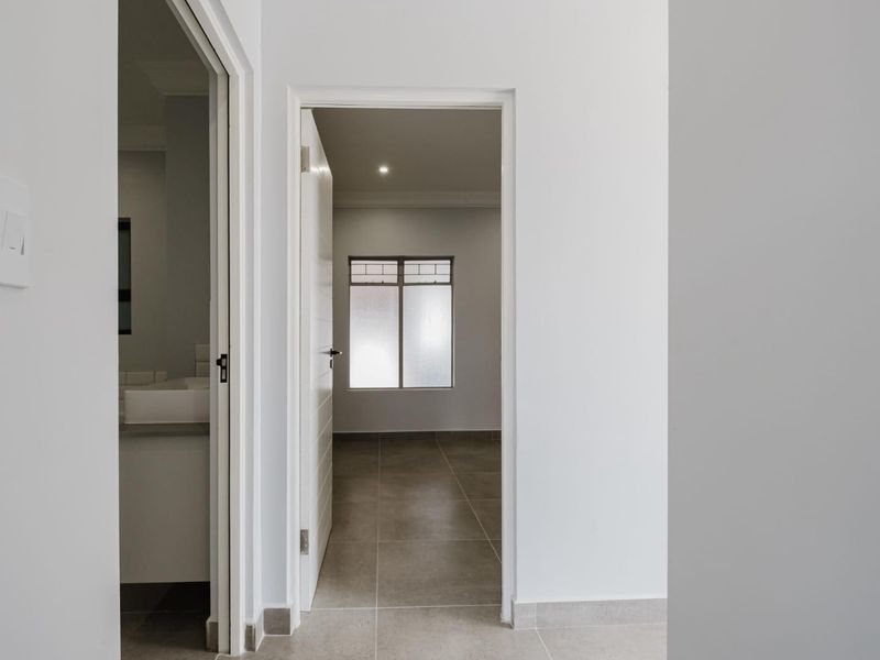 A clean, neutral hallway featuring tiled flooring and recessed lighting, providing access to adjacent rooms with a view into a bright, empty bedroom.