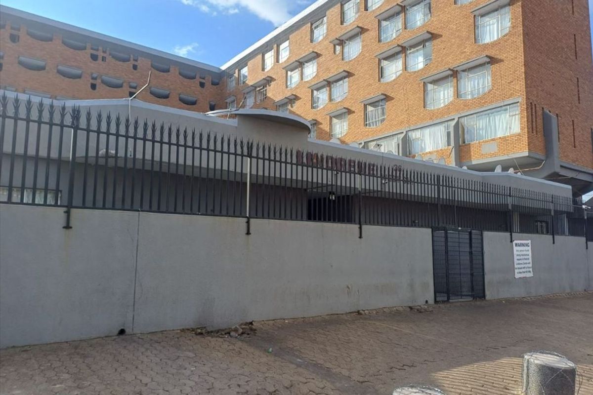 Exterior view of a multi-story brick apartment building featuring a secure perimeter wall, metal fencing, and paved driveway access.