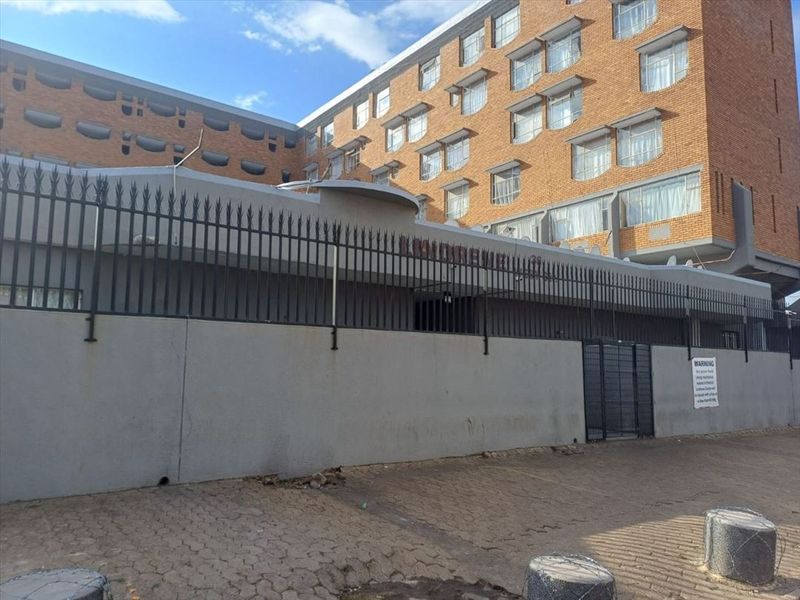 Exterior view of a multi-story brick apartment building featuring a secure perimeter wall, metal fencing, and paved driveway access.