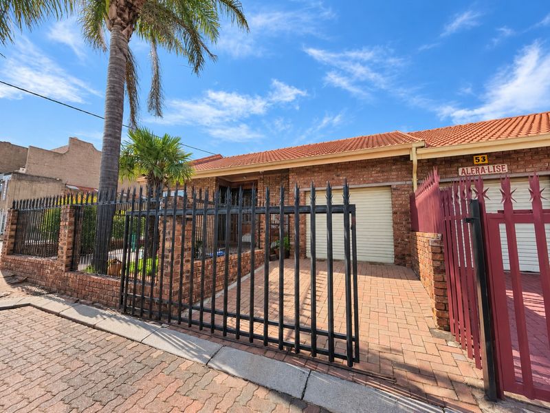 Front elevation of a brick house featuring a paved driveway, roller garage door, and secure metal perimeter fencing.