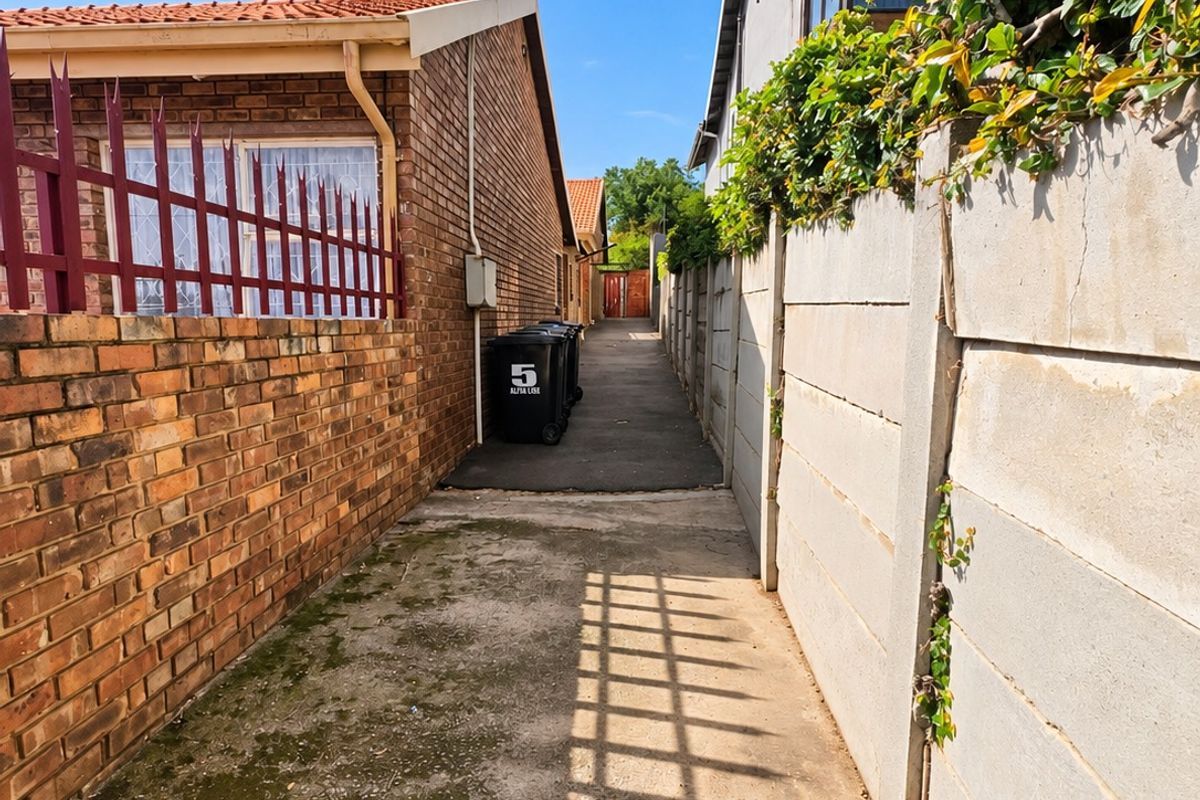A narrow paved side passage between two properties, featuring brick and precast concrete boundary walls with metal security fencing.