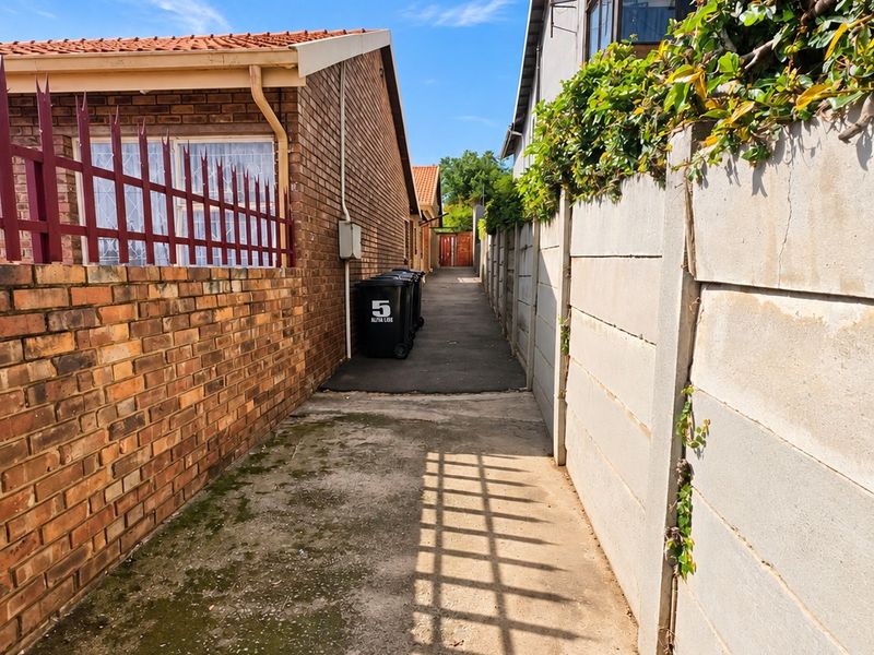 A narrow paved side passage between two properties, featuring brick and precast concrete boundary walls with metal security fencing.